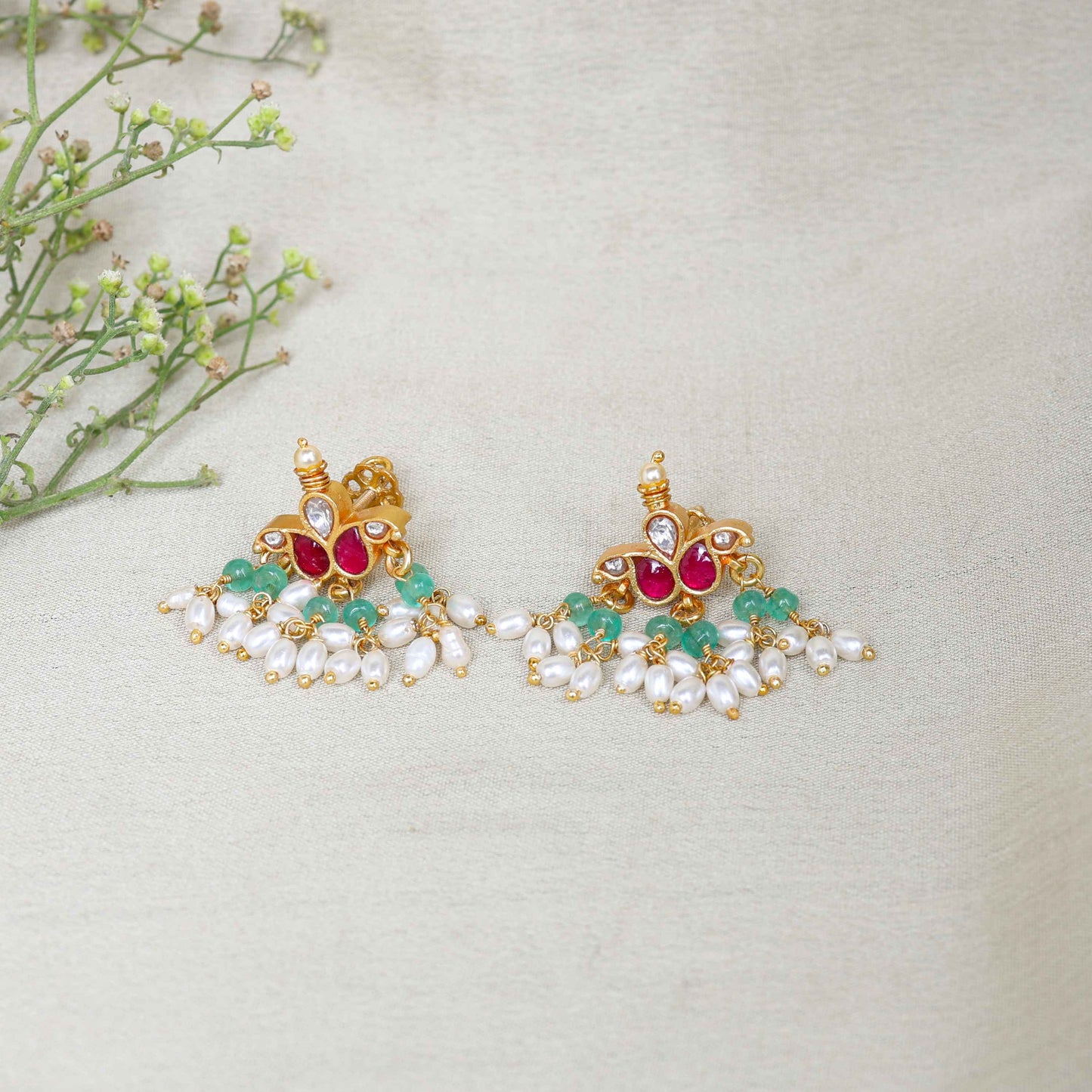 A pair of gold-plated Kundan chandelier earrings featuring ruby-red stones, emerald-green beads, and dangling freshwater pearls, displayed on a branch against a neutral background.