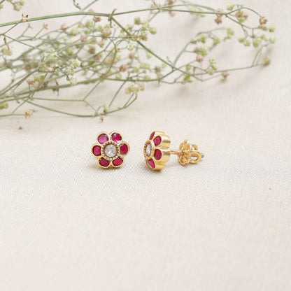 A close-up image of a woman's ear wearing a gold-toned floral stud earring, featuring a central white stone surrounded by pink gemstones, forming a flower shape.