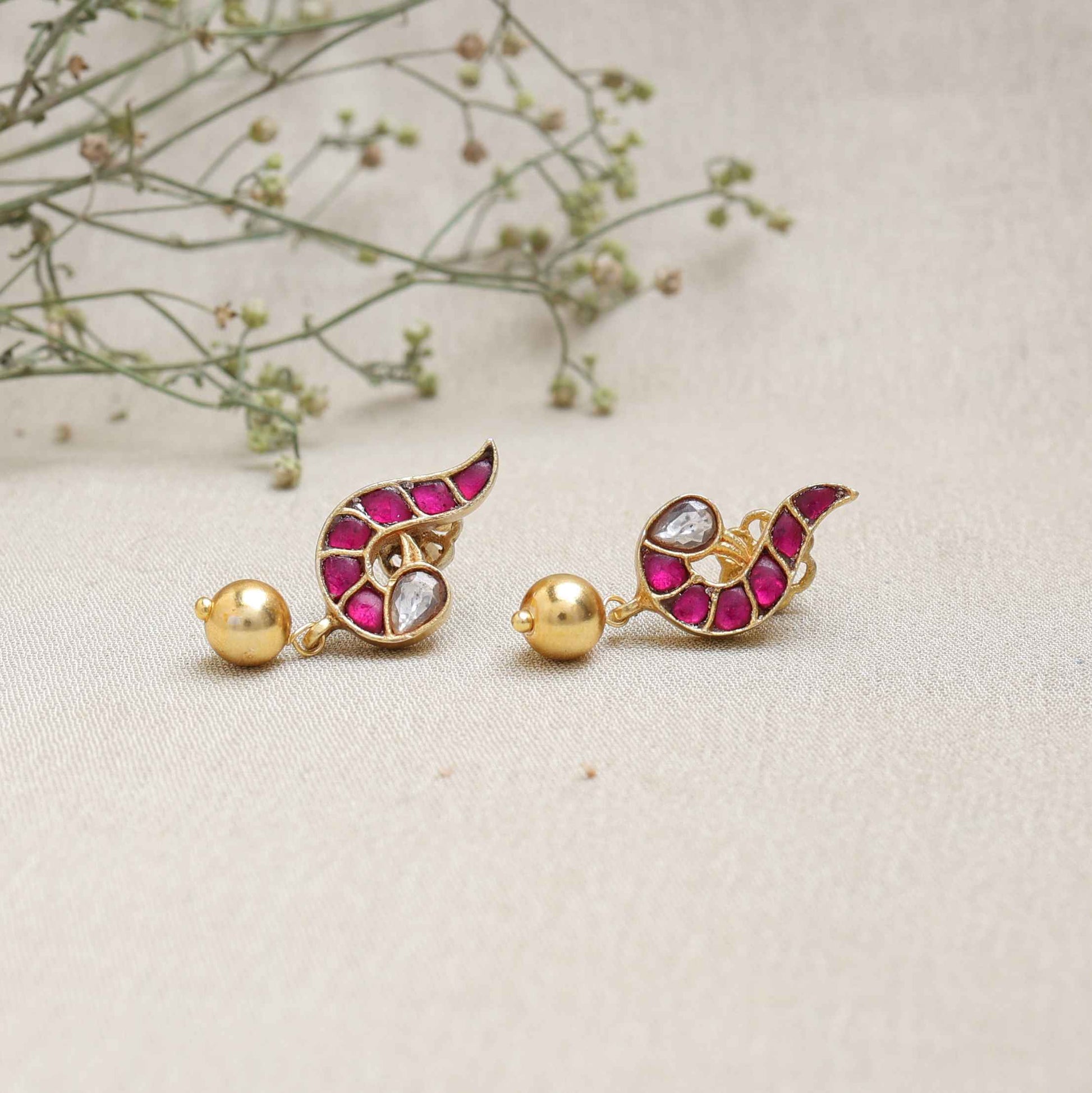 A close-up image of a woman's ear wearing a gold-toned paisley-shaped stud earring with ruby-red stones, a white gemstone accent, and a dangling gold bead.