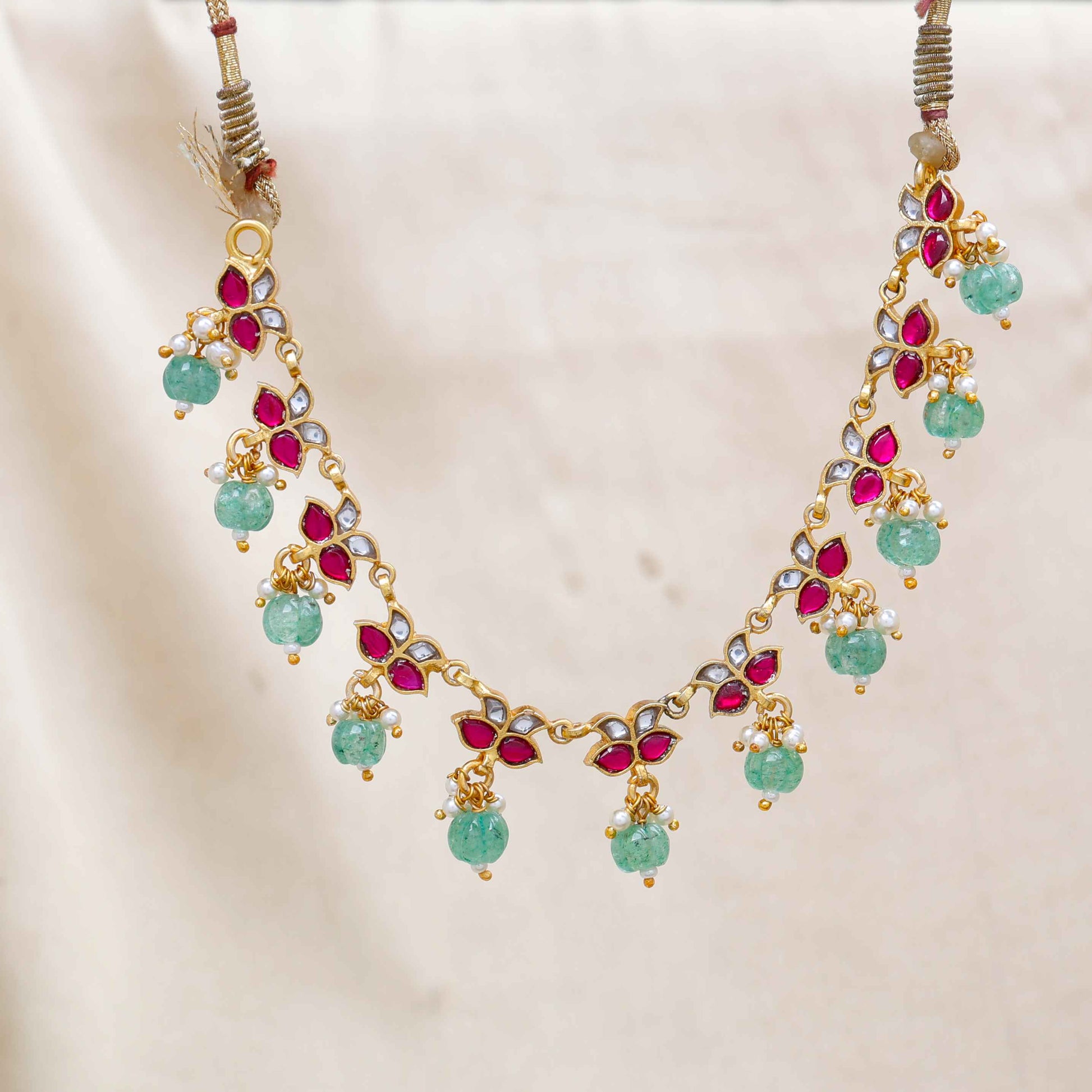 Gold-plated Kundan choker necklace with ruby-red, white stones, and dangling emerald-green beads, displayed on a neutral background.