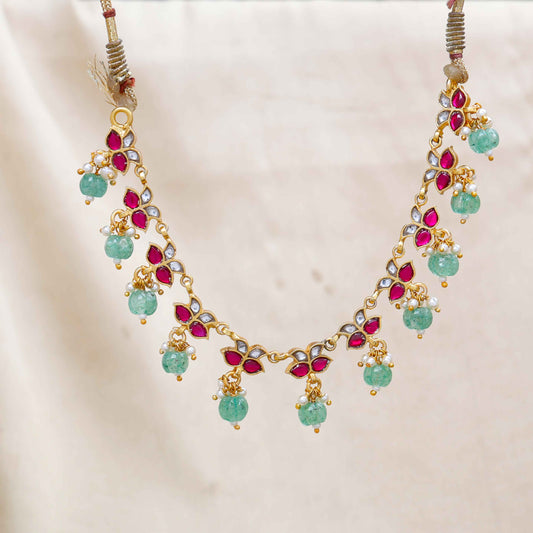 Gold-plated Kundan choker necklace with ruby-red, white stones, and dangling emerald-green beads, displayed on a neutral background.