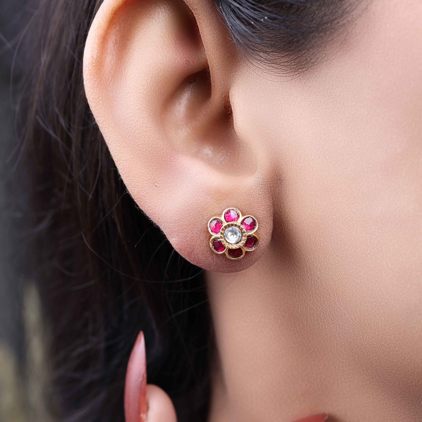 A close-up image of a woman's ear wearing a gold-toned floral stud earring, featuring a central white stone surrounded by pink gemstones, forming a flower shape.