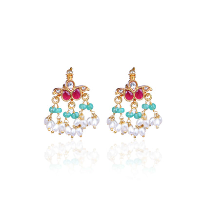 A pair of gold-plated Kundan chandelier earrings featuring ruby-red stones, emerald-green beads, and dangling freshwater pearls, displayed on a branch against a neutral background.