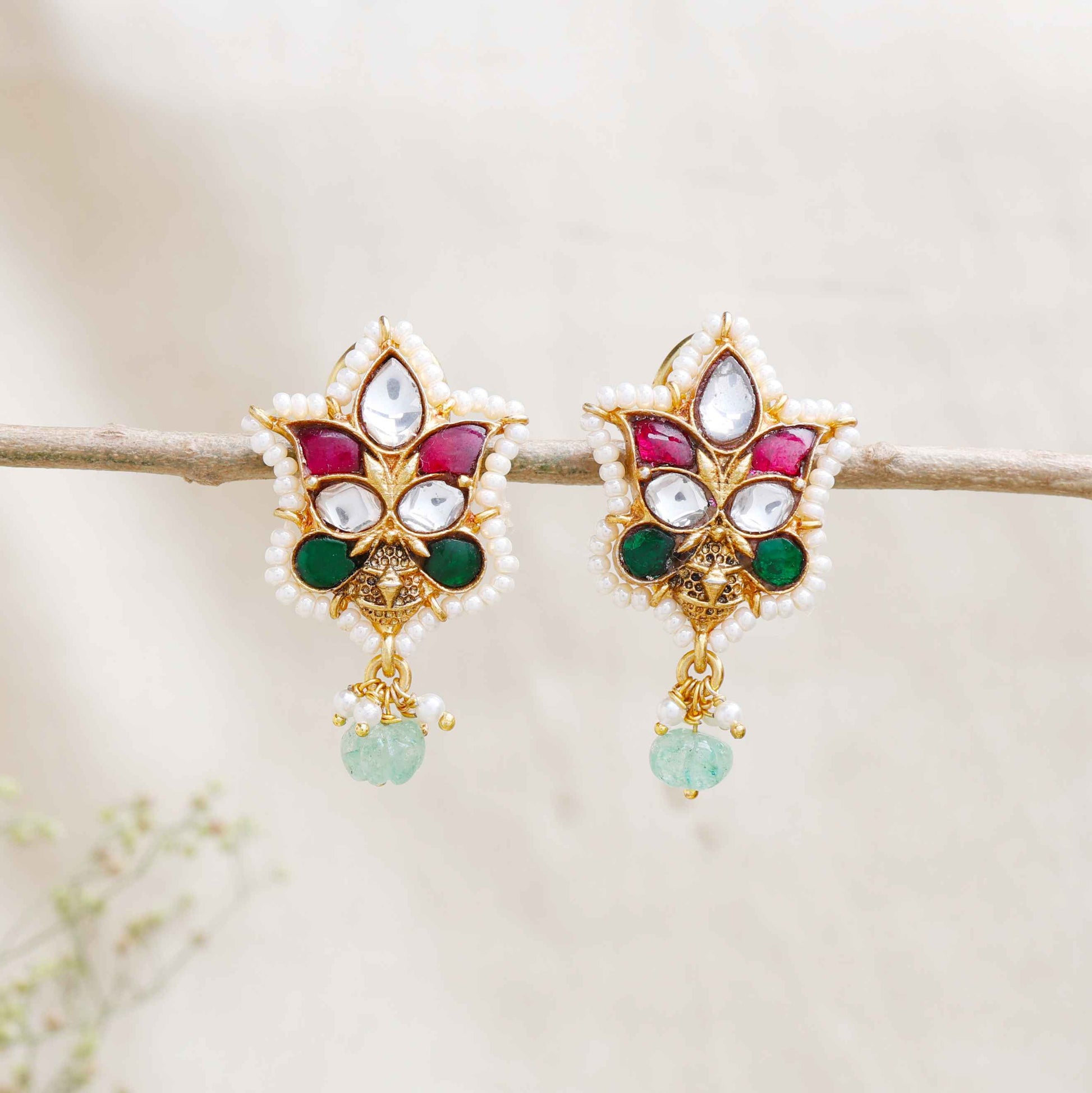 A close-up image of a woman's ear wearing a gold-toned Kundan stud earring with a floral design, featuring red, green, and white gemstones, surrounded by tiny pearls, and a dangling green bead at the bottom.