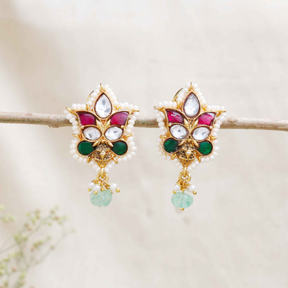 A close-up image of a woman's ear wearing a gold-toned Kundan stud earring with a floral design, featuring red, green, and white gemstones, surrounded by tiny pearls, and a dangling green bead at the bottom.