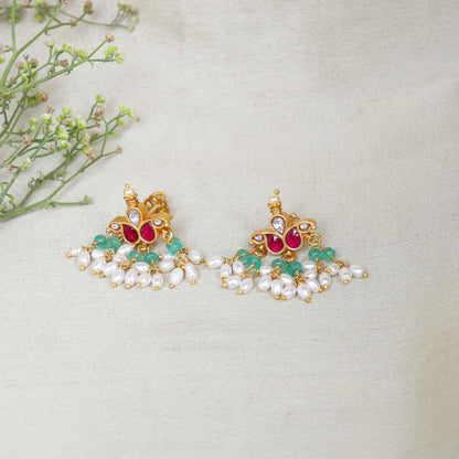 A pair of gold-plated Kundan chandelier earrings featuring ruby-red stones, emerald-green beads, and dangling freshwater pearls, displayed on a branch against a neutral background.