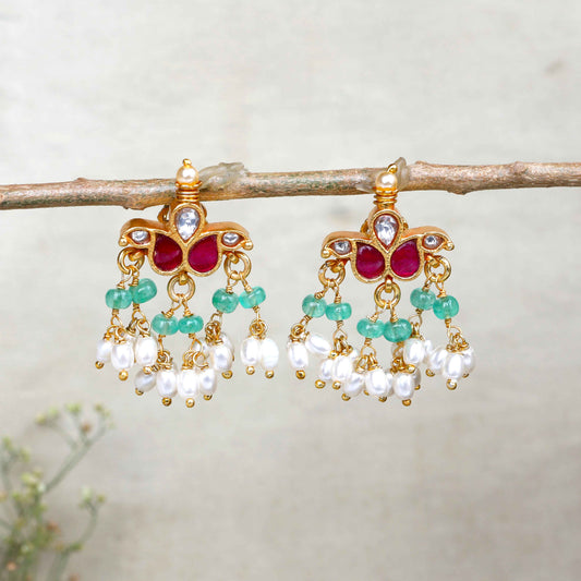 A pair of gold-plated Kundan chandelier earrings featuring ruby-red stones, emerald-green beads, and dangling freshwater pearls, displayed on a branch against a neutral background.