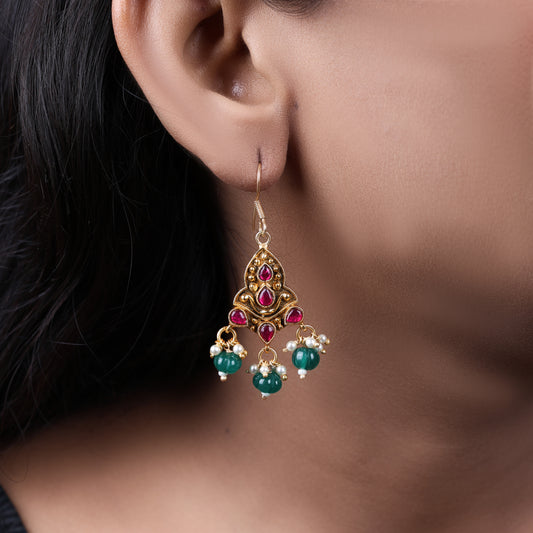 A close-up of a woman's ear adorned with a gold-toned chandelier-style earring featuring red teardrop-shaped stones, small pearl accents, and dangling green beads, exuding a traditional and elegant look.