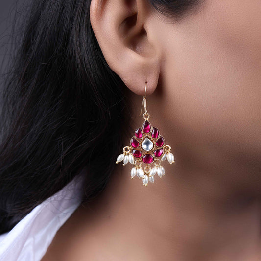 A close-up image of a woman's ear wearing a teardrop-shaped gold-toned drop earring, adorned with deep pink gemstones, a central white stone, and dangling white pearl-like beads.