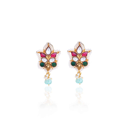 A close-up image of a woman's ear wearing a gold-toned Kundan stud earring with a floral design, featuring red, green, and white gemstones, surrounded by tiny pearls, and a dangling green bead at the bottom.