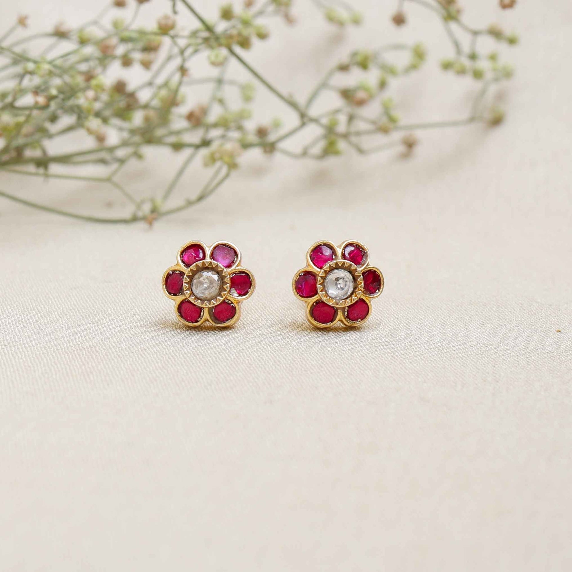 A close-up image of a woman's ear wearing a gold-toned floral stud earring, featuring a central white stone surrounded by pink gemstones, forming a flower shape.