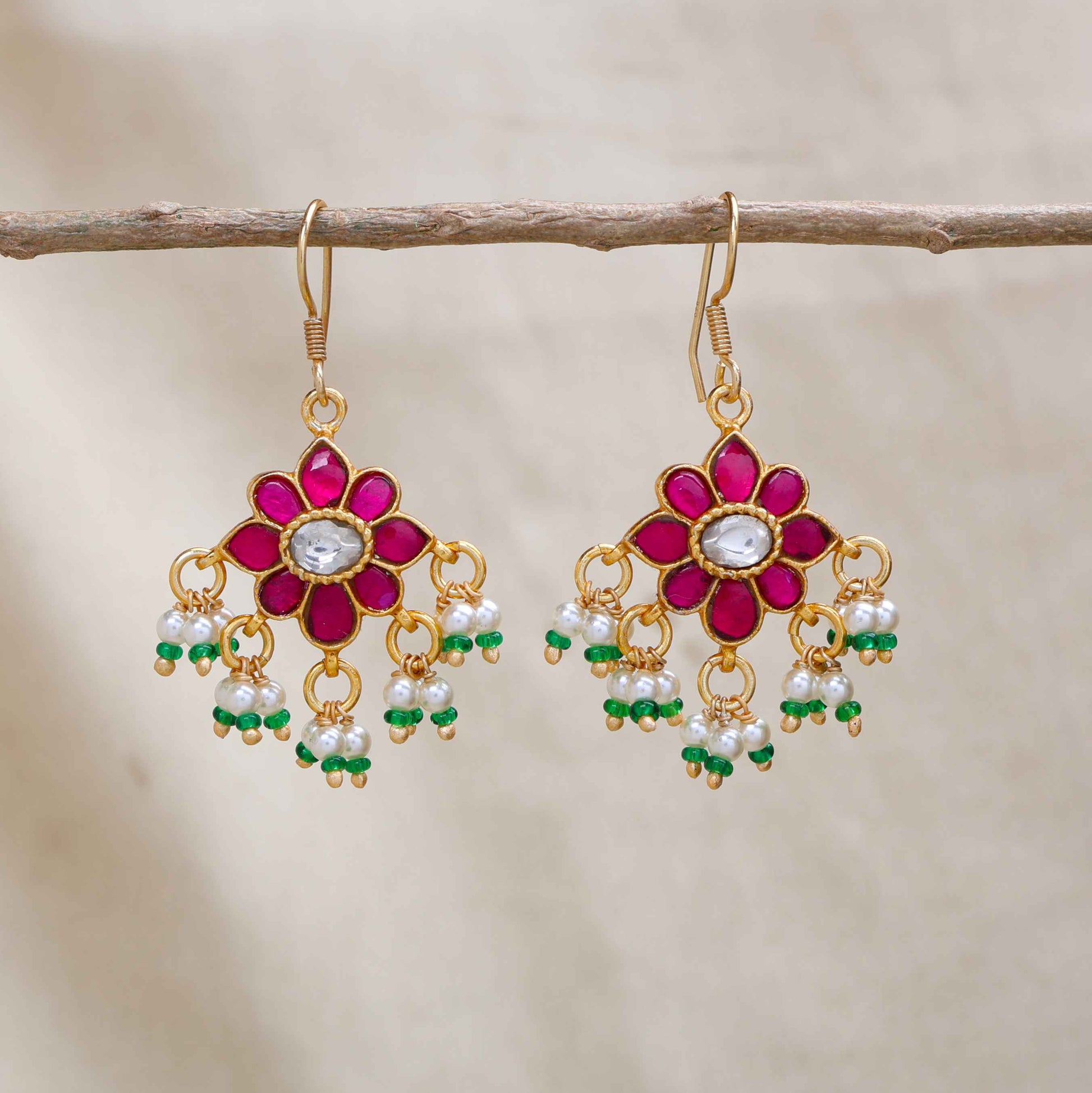 Gold-plated floral Kundan earrings with a central clear gemstone, surrounded by ruby-red stones and dangling pearl beads, worn by a model.