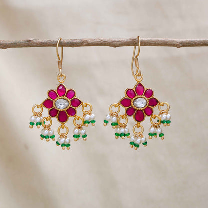 Gold-plated floral Kundan earrings with a central clear gemstone, surrounded by ruby-red stones and dangling pearl beads, worn by a model.