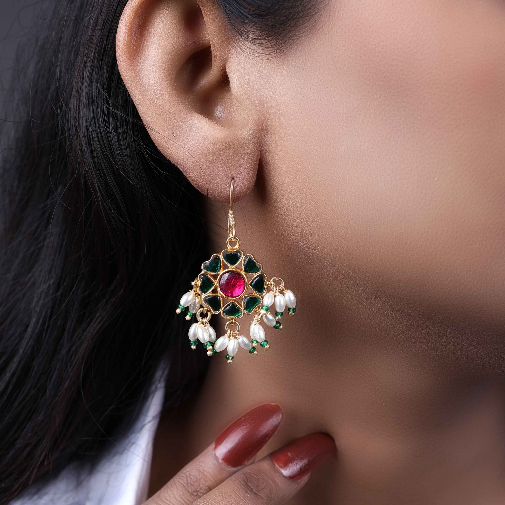 A gold-plated floral Kundan earring with a ruby-red center stone, deep green enamel petals, and dangling freshwater pearls, worn by a model with elegant styling.