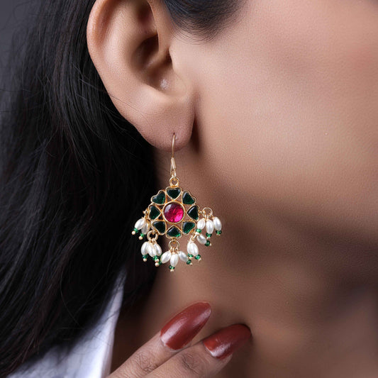 A gold-plated floral Kundan earring with a ruby-red center stone, deep green enamel petals, and dangling freshwater pearls, worn by a model with elegant styling.