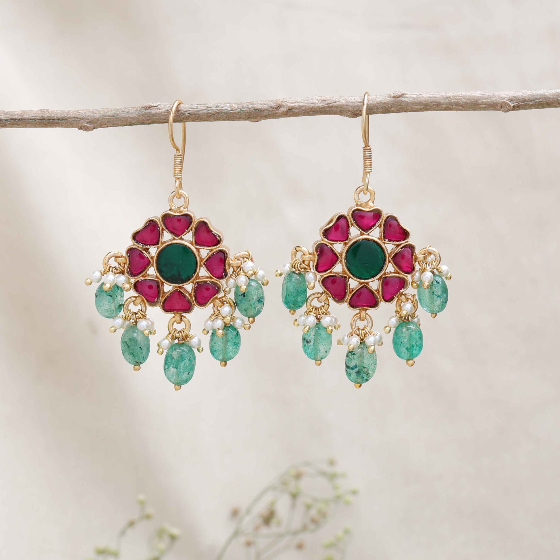 Elegant gold-plated Kundan earrings with a floral design, featuring a central green gemstone, ruby-red stones, and dangling emerald-green beads and pearls, worn by a model.
