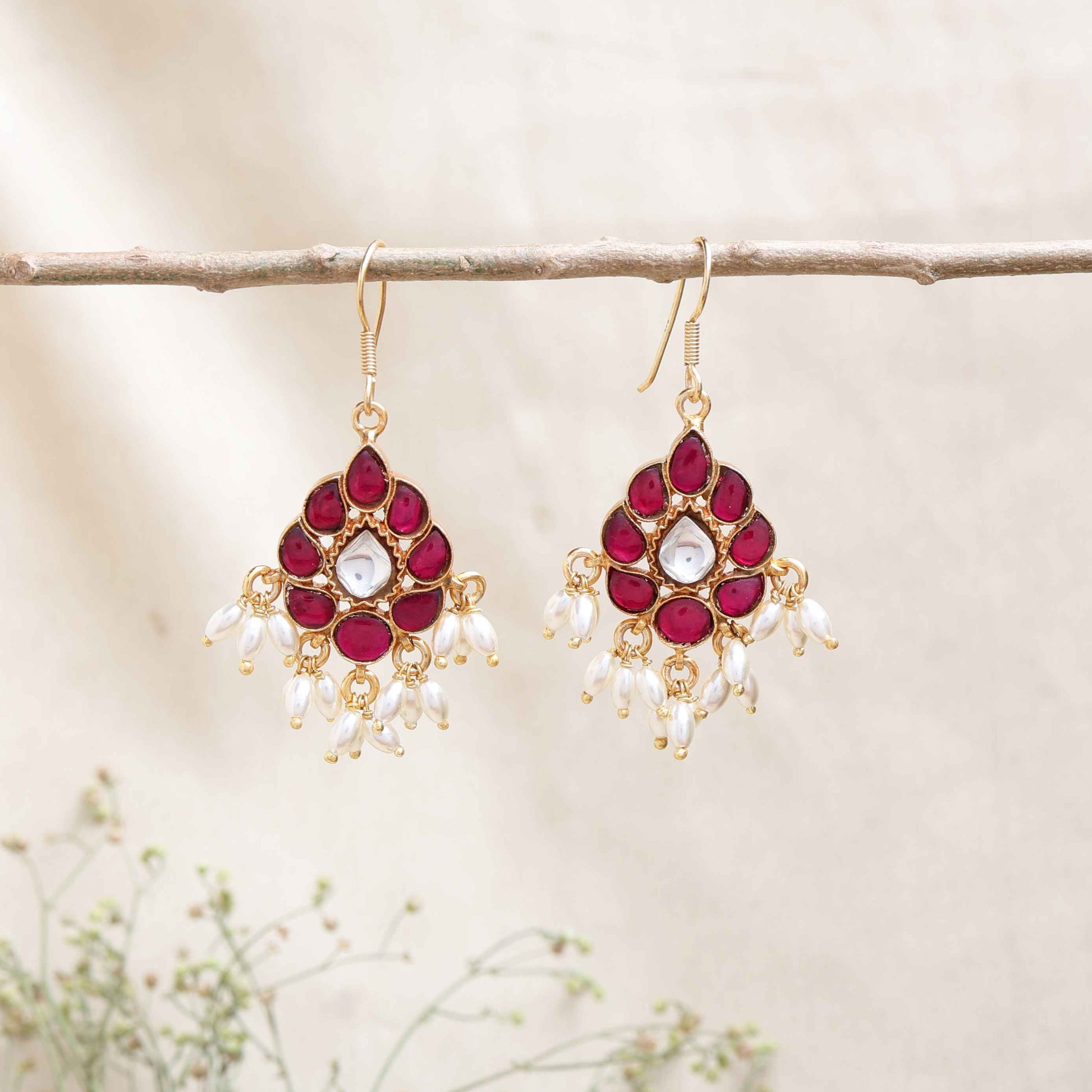 A close-up image of a woman's ear wearing a teardrop-shaped gold-toned drop earring, adorned with deep pink gemstones, a central white stone, and dangling white pearl-like beads.