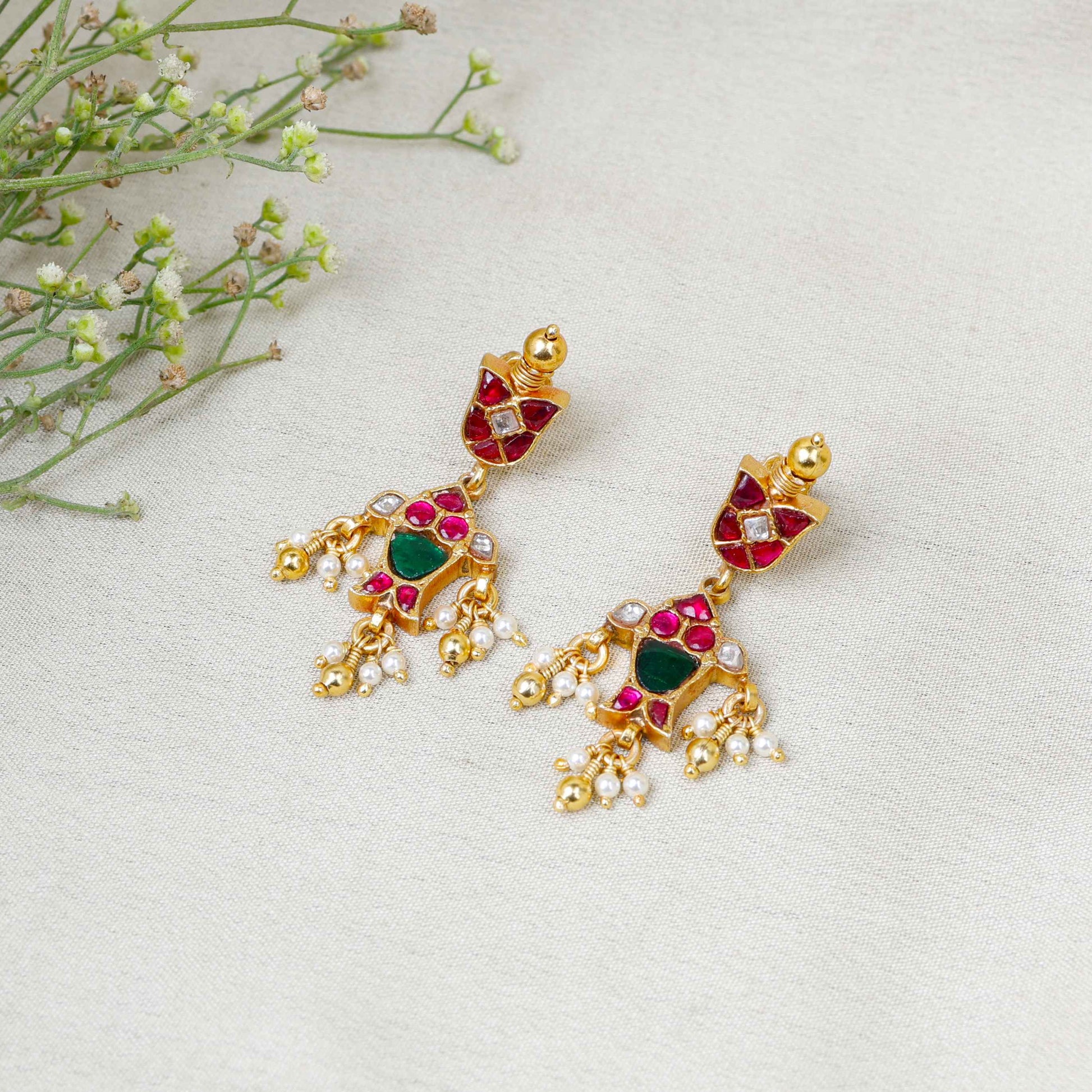 Gold-plated Kundan earrings with red and green gemstones, intricate floral design, and dangling pearl drops, perfect for traditional and festive wear.