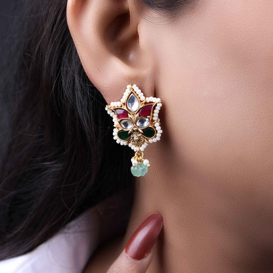 A close-up image of a woman's ear wearing a gold-toned Kundan stud earring with a floral design, featuring red, green, and white gemstones, surrounded by tiny pearls, and a dangling green bead at the bottom.