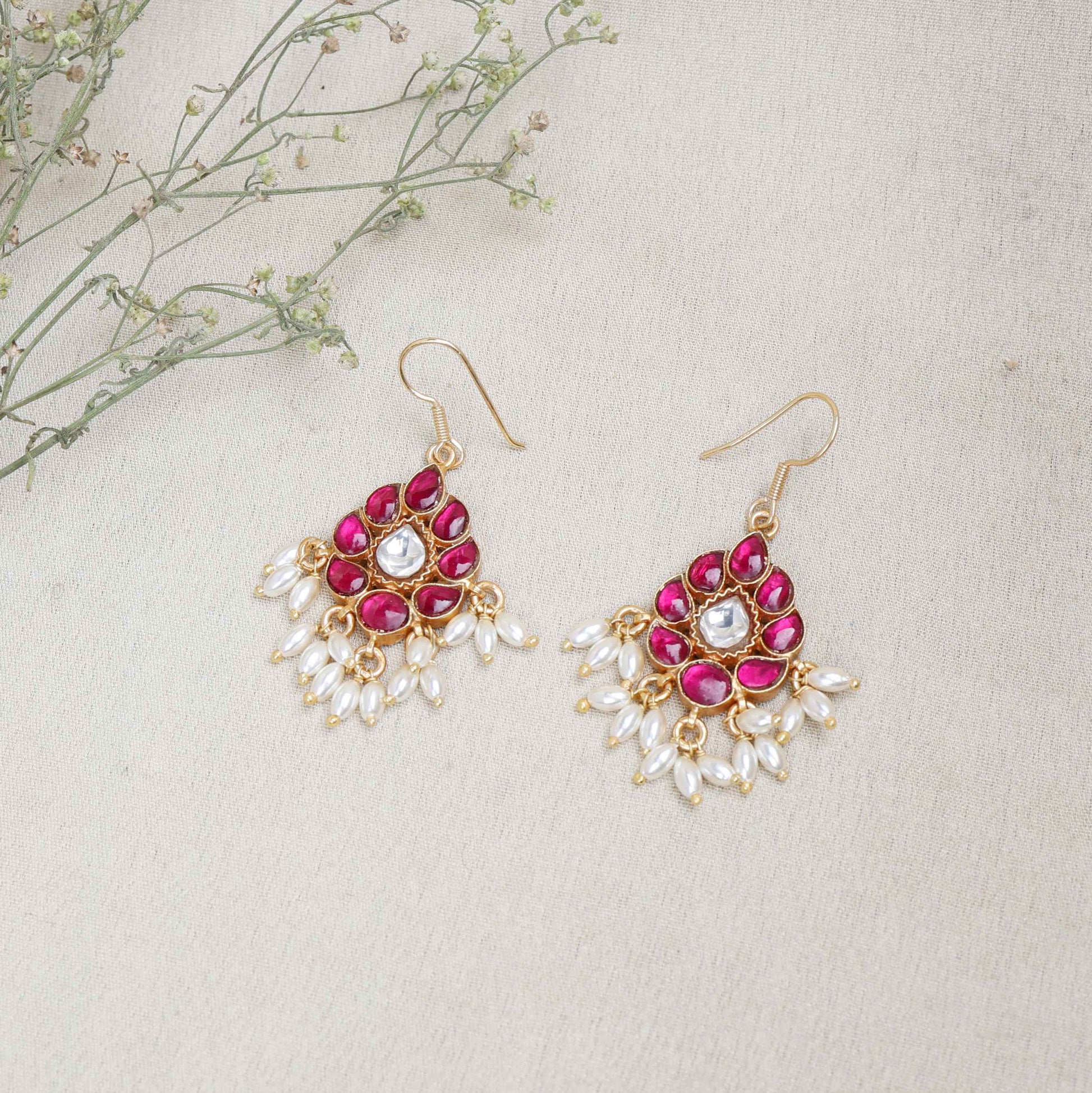 A close-up image of a woman's ear wearing a teardrop-shaped gold-toned drop earring, adorned with deep pink gemstones, a central white stone, and dangling white pearl-like beads.