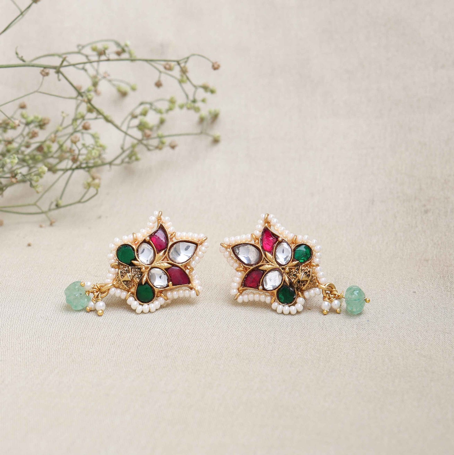 A close-up image of a woman's ear wearing a gold-toned Kundan stud earring with a floral design, featuring red, green, and white gemstones, surrounded by tiny pearls, and a dangling green bead at the bottom.