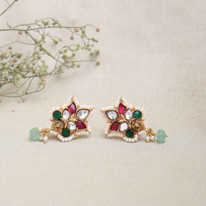 A close-up image of a woman's ear wearing a gold-toned Kundan stud earring with a floral design, featuring red, green, and white gemstones, surrounded by tiny pearls, and a dangling green bead at the bottom.