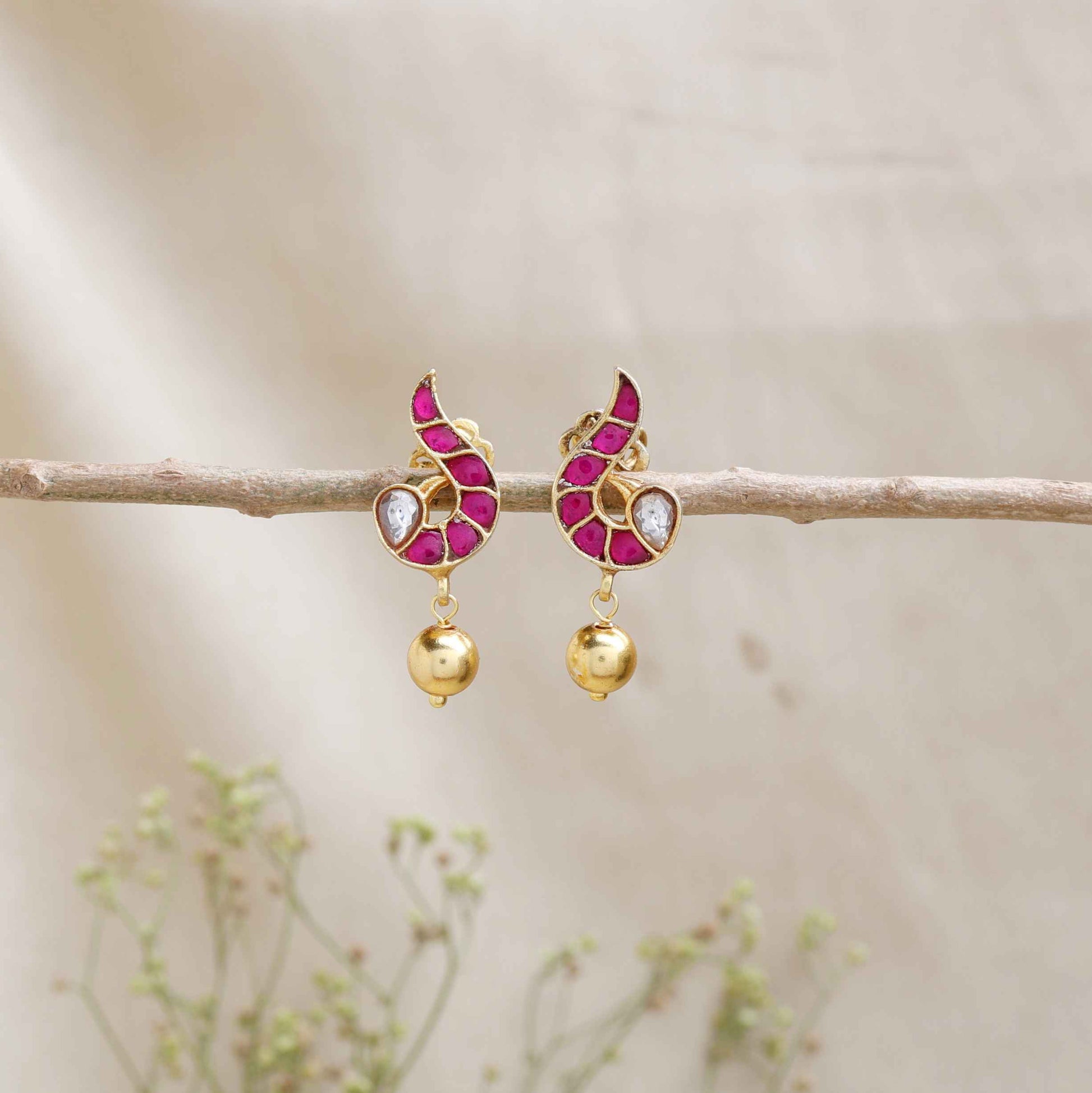 A close-up image of a woman's ear wearing a gold-toned paisley-shaped stud earring with ruby-red stones, a white gemstone accent, and a dangling gold bead.