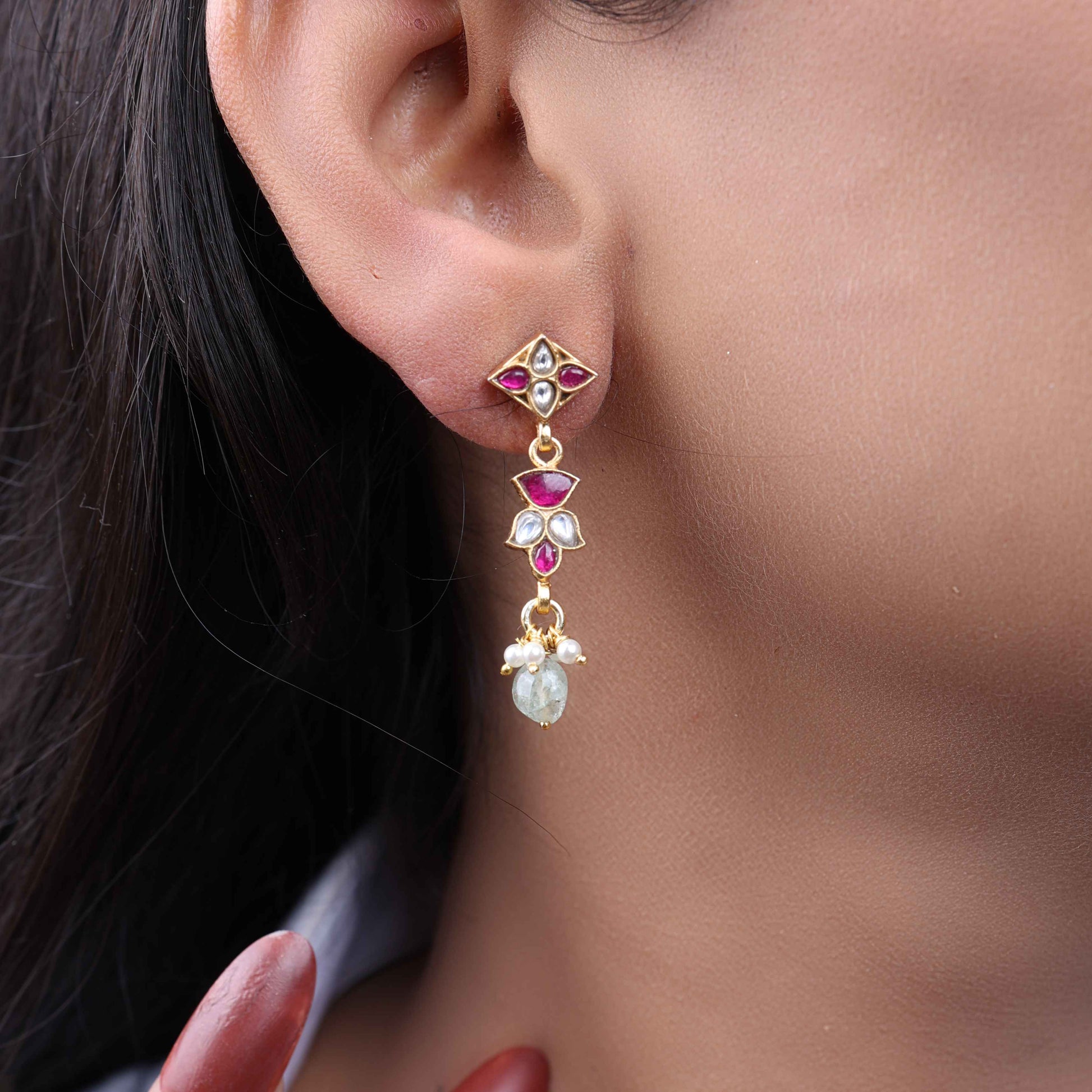A close-up image of a woman's ear wearing gold-toned drop earrings with ruby-red and white stones in a geometric design, accented with pearls and a dangling light green gemstone.