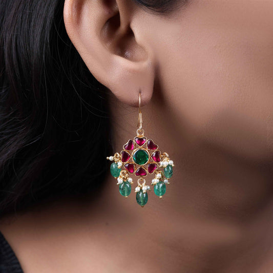 Elegant gold-plated Kundan earrings with a floral design, featuring a central green gemstone, ruby-red stones, and dangling emerald-green beads and pearls, worn by a model.