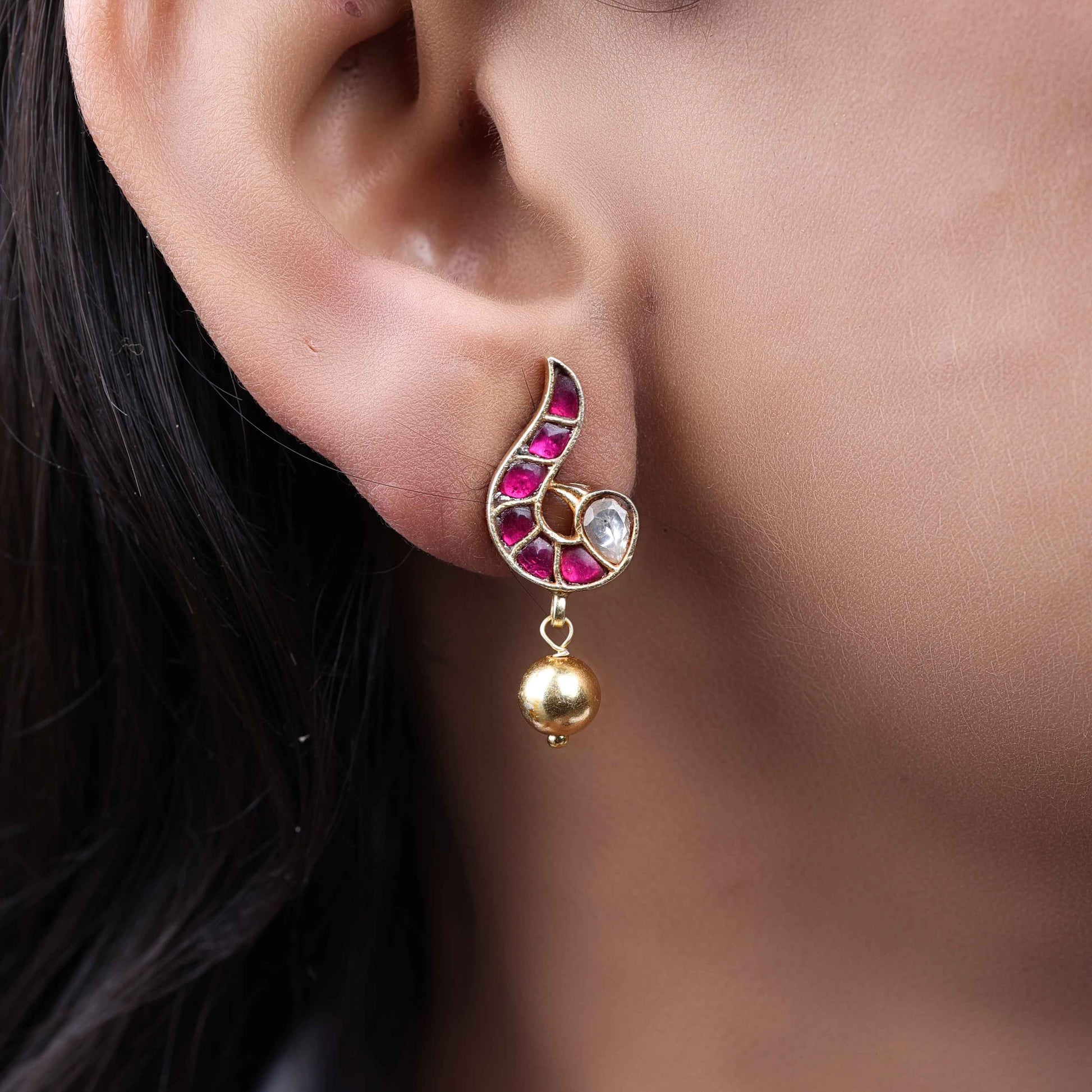 A close-up image of a woman's ear wearing a gold-toned paisley-shaped stud earring with ruby-red stones, a white gemstone accent, and a dangling gold bead.