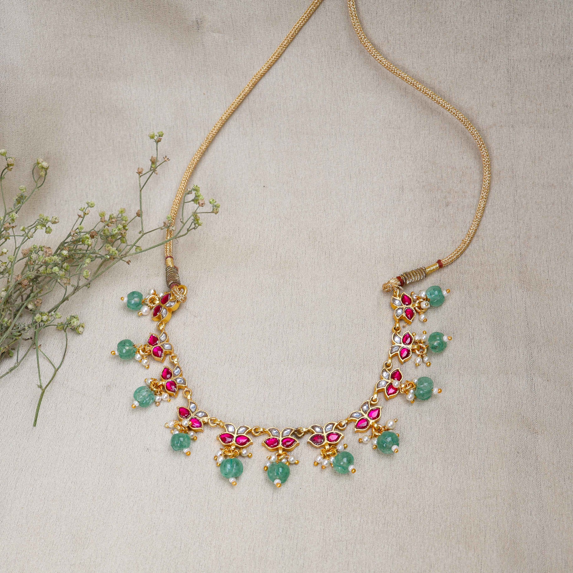 Gold-plated Kundan choker necklace with ruby-red, white stones, and dangling emerald-green beads, displayed on a neutral background.