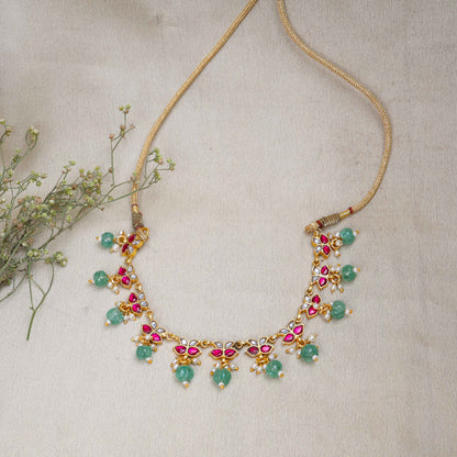 Gold-plated Kundan choker necklace with ruby-red, white stones, and dangling emerald-green beads, displayed on a neutral background.