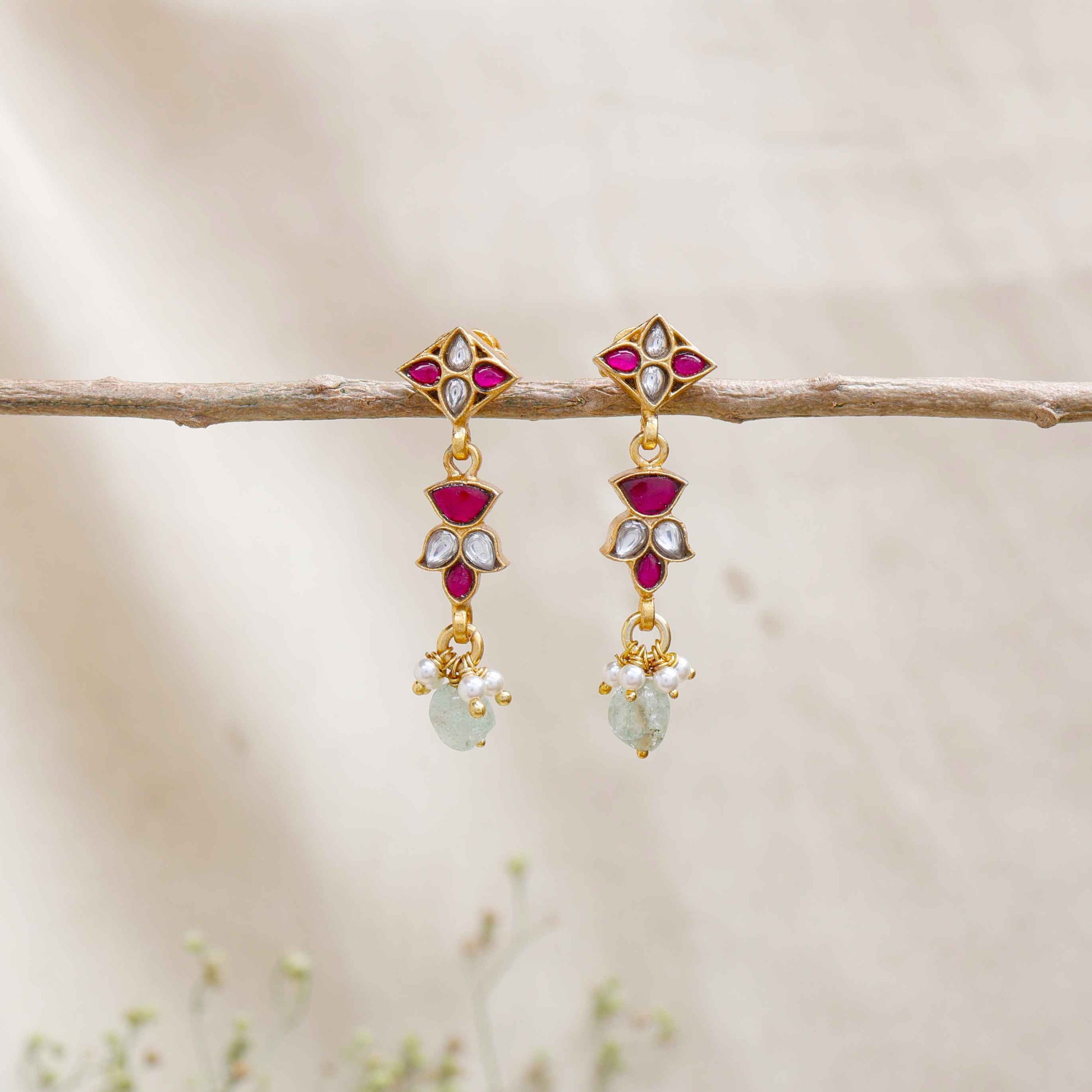 A close-up image of a woman's ear wearing gold-toned drop earrings with ruby-red and white stones in a geometric design, accented with pearls and a dangling light green gemstone.