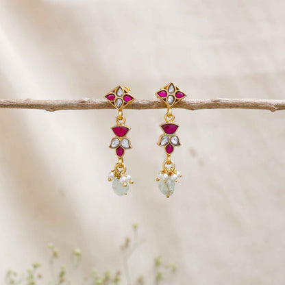 A close-up image of a woman's ear wearing gold-toned drop earrings with ruby-red and white stones in a geometric design, accented with pearls and a dangling light green gemstone.