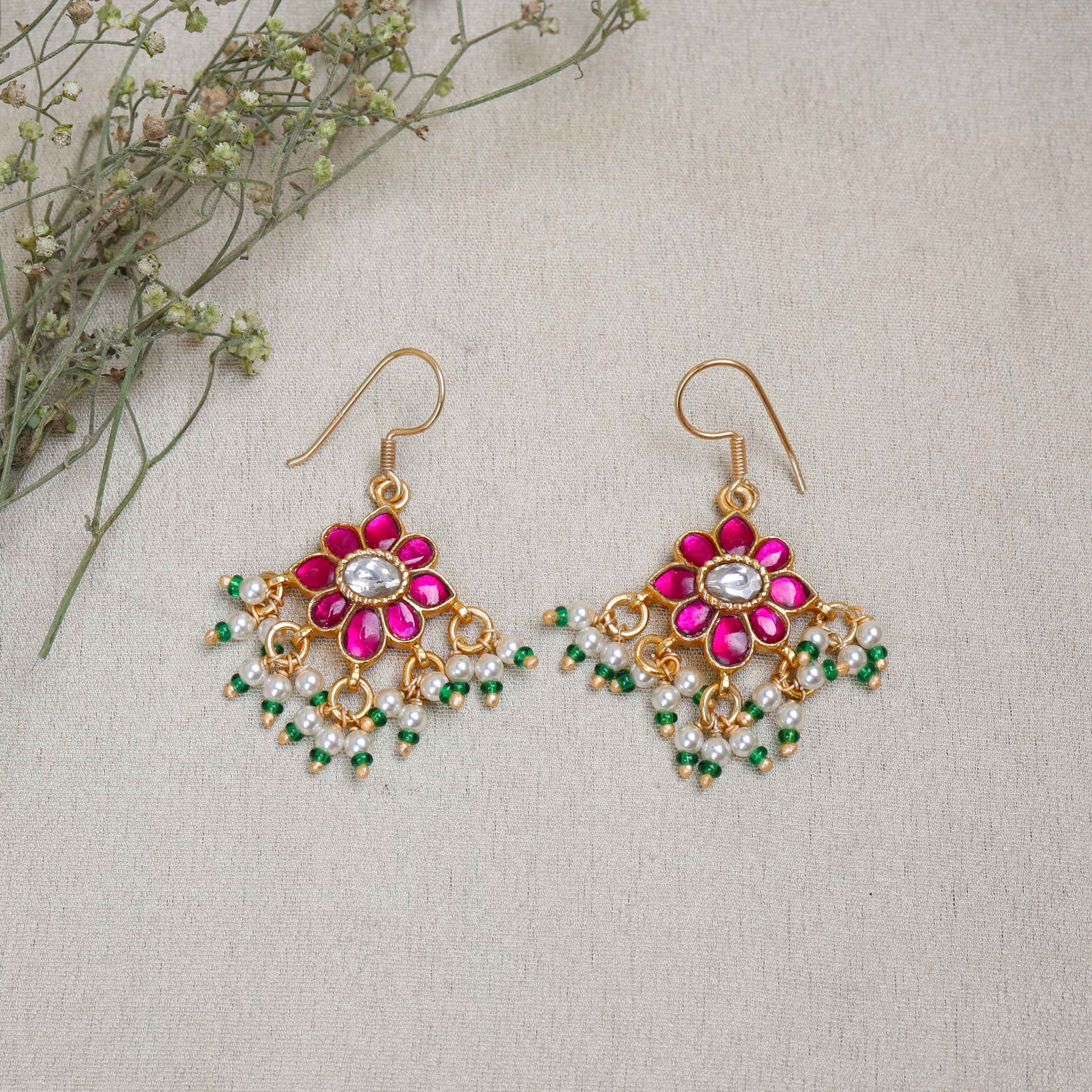 Gold-plated floral Kundan earrings with a central clear gemstone, surrounded by ruby-red stones and dangling pearl beads, worn by a model.