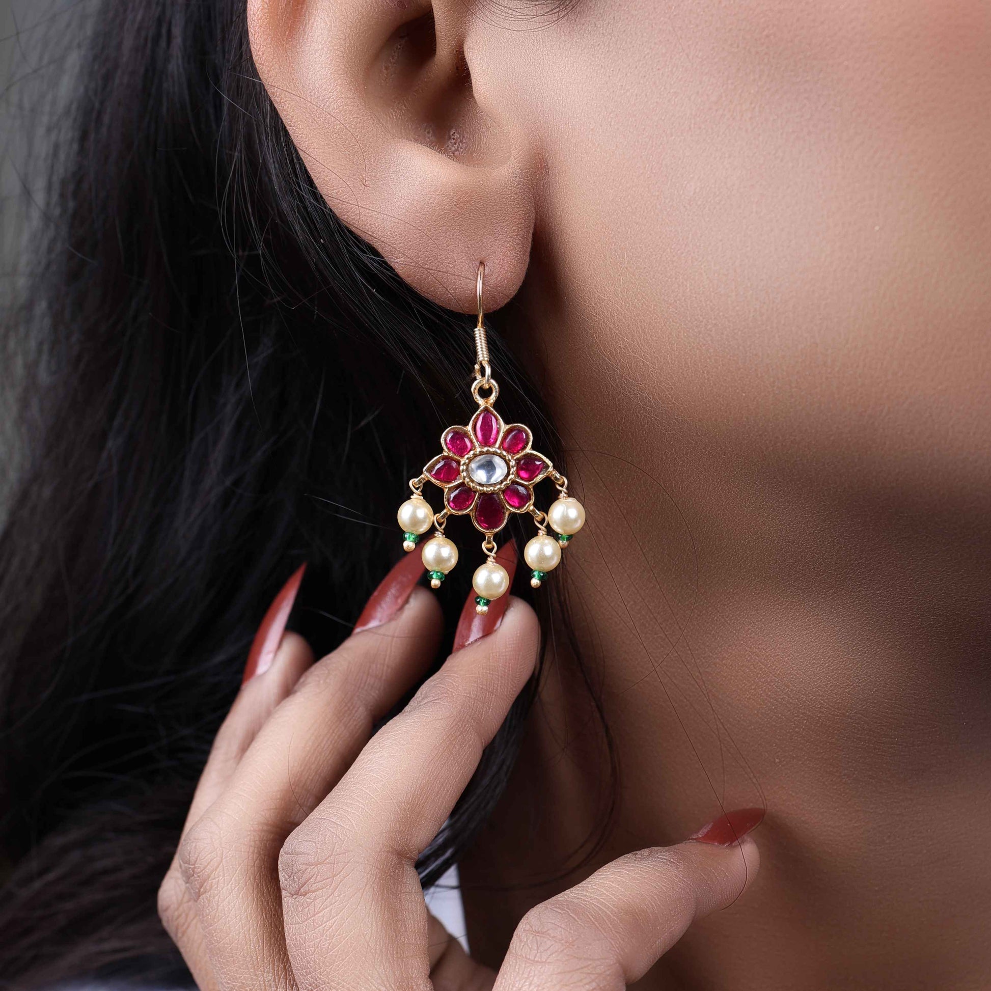 Gold-plated floral Kundan earrings with a central clear gemstone, surrounded by ruby-red stones and dangling pearl beads, worn by a model.