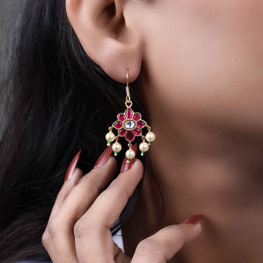 Gold-plated floral Kundan earrings with a central clear gemstone, surrounded by ruby-red stones and dangling pearl beads, worn by a model.