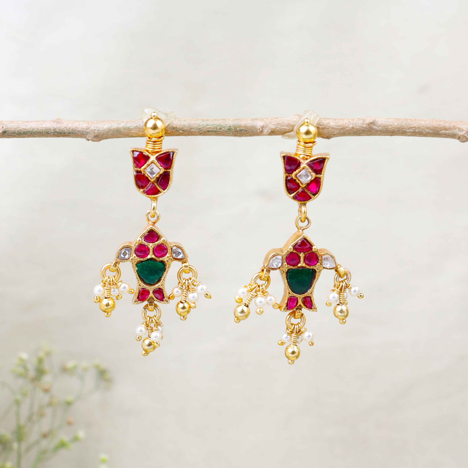 Gold-plated Kundan earrings with red and green gemstones, intricate floral design, and dangling pearl drops, perfect for traditional and festive wear.